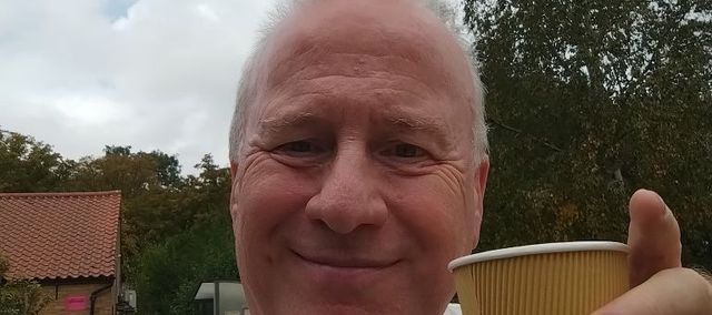 Nigel sitting in a country park smiling and holding up a beige coffee mug in salute.